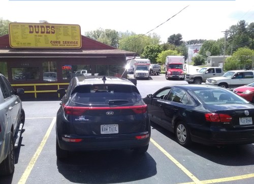 A parking lot with two cars in front of a restaurant called "Dudes" and a U-Haul truck in the background.