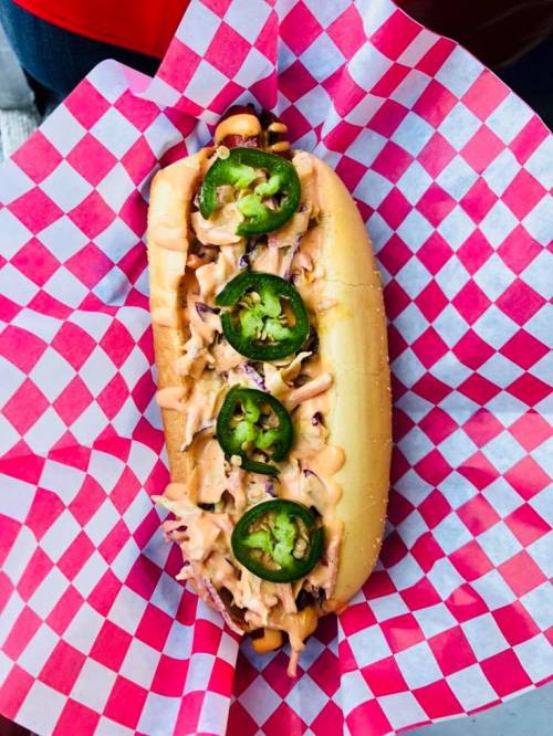 A hot dog topped with sliced jalapeños and a creamy sauce, served in a checkered paper basket.