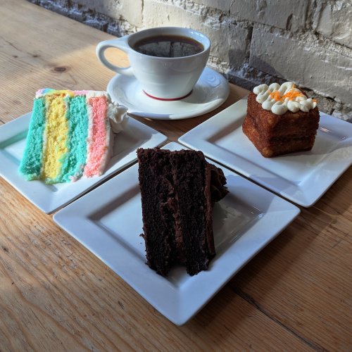A cup of coffee beside three dessert slices: rainbow cake, chocolate cake, and a frosted square cake on white plates.
