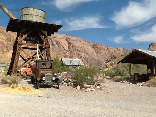 An old truck and rustic buildings under a blue sky, surrounded by rocky hills and desert vegetation.