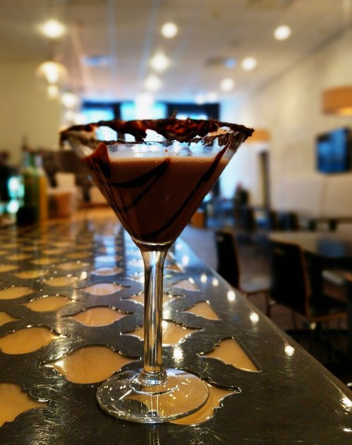 A chocolate martini with a decorative rim sits on a bar counter, with a blurred background of a modern lounge.