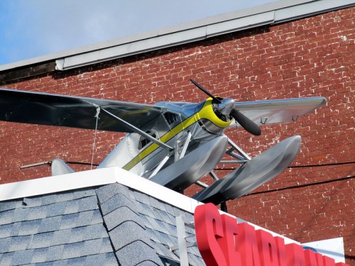 A vintage silver airplane with a yellow stripe perched on the roof of a building against a blue sky.