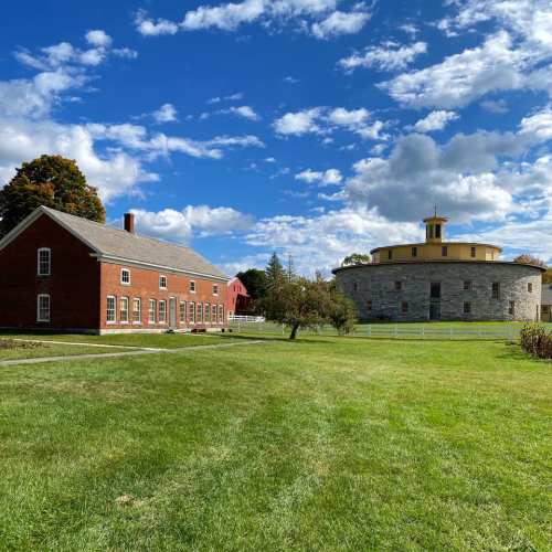 A scenic view of a grassy area featuring a red brick building, a round stone structure, and a blue sky with clouds.