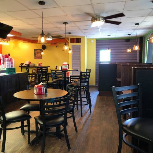 A brightly lit restaurant interior with wooden floors, round tables, and black chairs, featuring pendant lights and a counter.