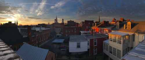 Panoramic view of a town at sunset, featuring rooftops, steeples, and a colorful sky.