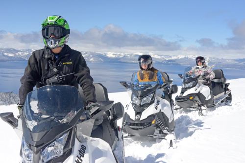 Three snowmobilers on a snowy mountain trail with a lake and mountains in the background under a clear blue sky.