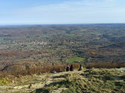 A group of people sits on a rocky ledge, overlooking a vast valley with autumn foliage and a small town below.