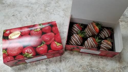 A box of chocolate-covered strawberries, some drizzled with white chocolate, next to a decorated box featuring strawberries.