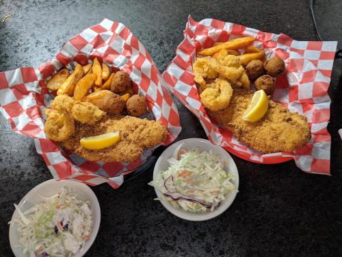 Two baskets of fried seafood with lemon, sweet potato fries, hushpuppies, and bowls of coleslaw on a black surface.
