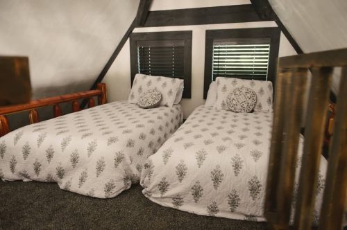 Cozy attic bedroom with two beds, white bedding, and black-framed windows. Rustic wooden accents visible.