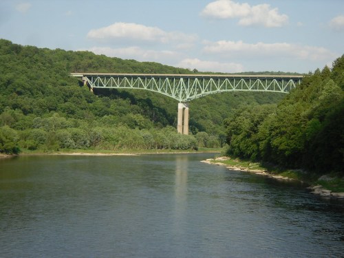A green steel bridge spans a river, surrounded by lush green trees and hills under a partly cloudy sky.