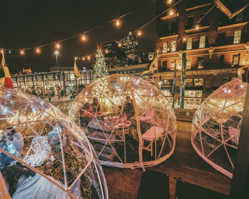 Cozy outdoor dining igloos illuminated with string lights, set against a city skyline at night.