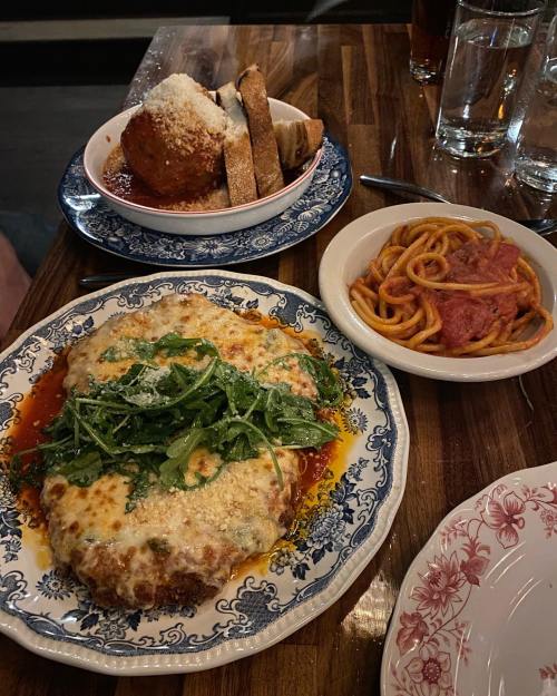 A plate of chicken parmesan topped with arugula, a bowl of spaghetti with meat, and a side of meatballs with bread.