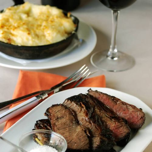 A plate of sliced grilled steak with a side of salt, accompanied by a glass of red wine and a dish of cheesy potatoes.