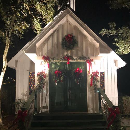 A charming white chapel decorated with wreaths and red bows, illuminated at night with festive lights.