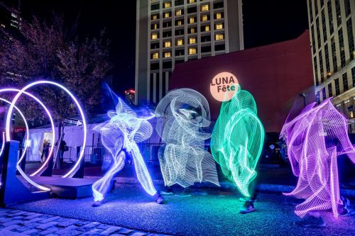 Colorful light figures dance in a city square during the LUNA Fête, surrounded by illuminated structures.
