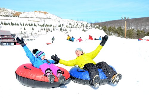 Two people in colorful winter gear joyfully sit in snow tubes, surrounded by a snowy landscape and other tubers in the background.