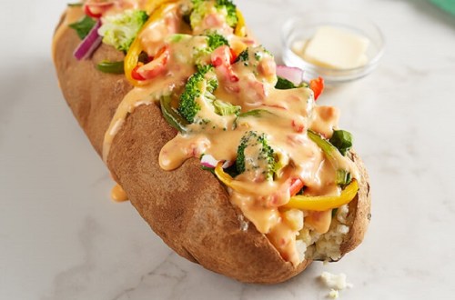 A loaded baked potato topped with melted cheese and colorful vegetables, served with a small dish of butter.