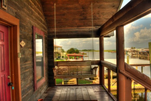 A wooden porch with a swing overlooks a calm waterway and nearby houses under a cloudy sky.
