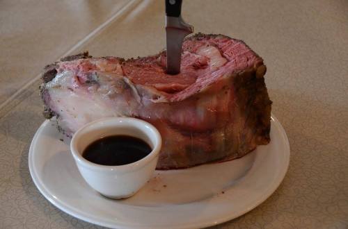 A large cut of roast beef on a plate with a knife inserted and a small bowl of sauce beside it.