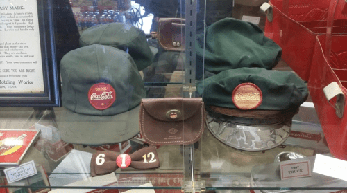 Display of vintage Coca-Cola hats and a leather pouch in a glass case, showcasing nostalgic memorabilia.