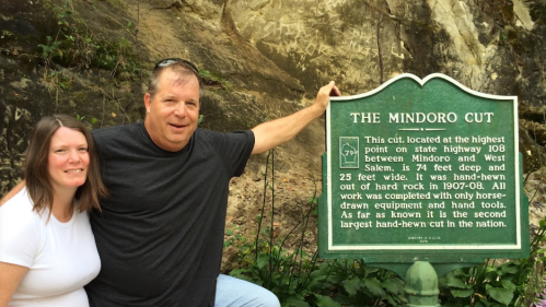 A man and woman pose beside a historical sign about the Mindoro Cut, detailing its dimensions and construction history.