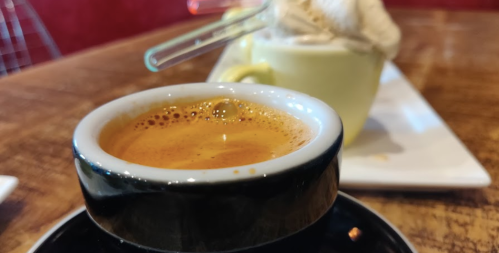 A close-up of a black espresso cup with rich crema, with a dessert in the background on a wooden table.