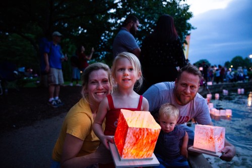 A family poses by a pond at dusk, holding glowing lanterns, with people and trees in the background.
