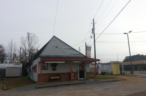 A small, single-story building with a peaked roof, surrounded by gravel and power lines, under a cloudy sky.
