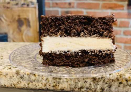 A slice of chocolate cake with a creamy filling, displayed on a clear plate against a brick background.