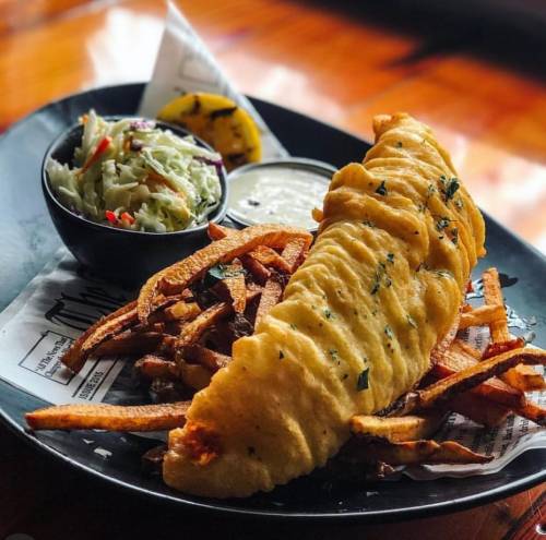 A plate featuring a large fried fish fillet, sweet potato fries, coleslaw, and a lemon wedge with dipping sauce.