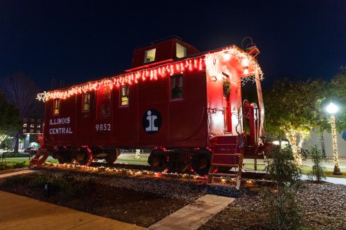 A red Illinois Central caboose decorated with festive lights, set against a night sky in a park.