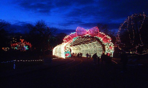 A festive archway adorned with colorful Christmas lights and a large bow, with people walking underneath at dusk.