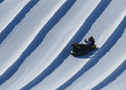 Two people sledding down a snowy slope with wavy patterns in the snow. Bright blue sky above.