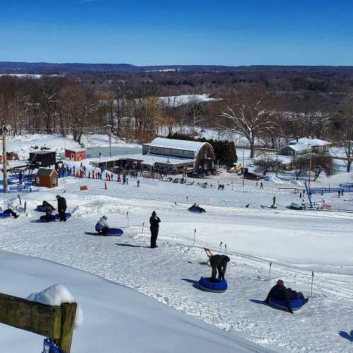 A snowy hill with people sledding down, surrounded by trees and a clear blue sky. A ski area is visible in the background.