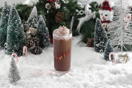 A festive chocolate drink topped with whipped cream, surrounded by snow, pine trees, and holiday decorations.