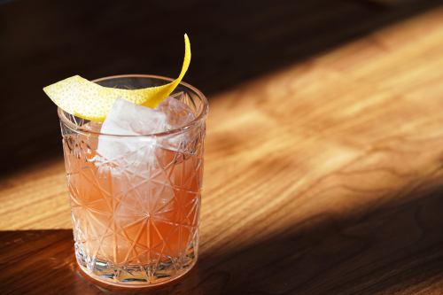 A glass of cocktail with ice and a lemon twist, resting on a wooden table with soft lighting.
