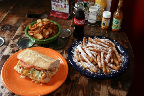 A sandwich with fried chicken and toppings, served with seasoned potato wedges and powdered sugar-coated fries, plus a Coke.