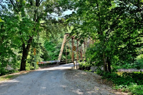 A gravel road leads to a rustic metal bridge surrounded by lush green trees and foliage.