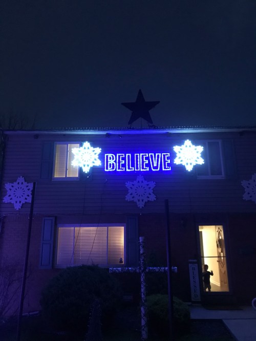 A house decorated with blue lights, featuring the word "BELIEVE" and snowflake designs against a dark evening sky.