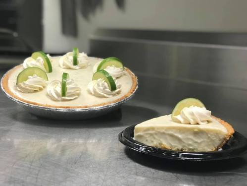 A whole key lime pie with whipped cream and lime slices, next to a slice on a black plate.