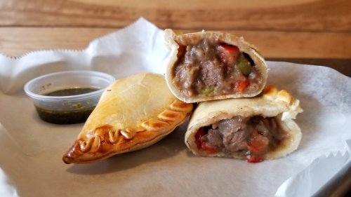 Two golden-brown empanadas filled with beef and vegetables, served with a small container of green sauce.