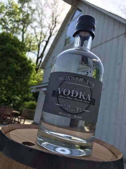 A clear vodka bottle with a black label sits on a wooden barrel, with a distillery building in the background.