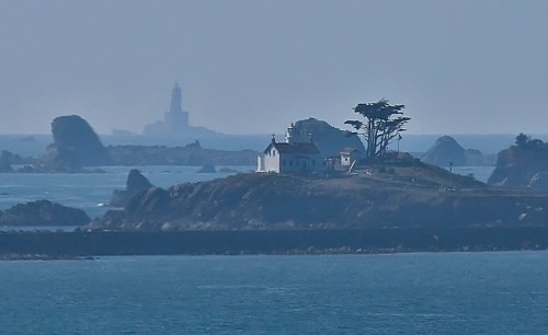 A distant view of a small island with a white building and trees, surrounded by rocky shores and a lighthouse in the background.