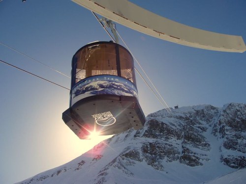 A gondola tram glides above snowy mountains, with sunlight reflecting off its surface against a clear blue sky.
