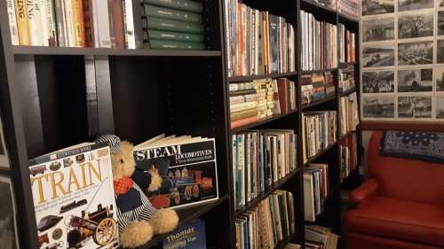 Bookshelves filled with various books, a teddy bear in a train outfit, and a red armchair in a cozy reading nook.