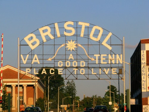 Sign archway reading "Bristol VA A Good Place to Live," with buildings and trees in the background under a clear sky.