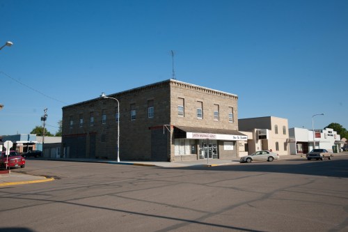 A two-story stone building on a street corner, with nearby shops and clear blue sky above.