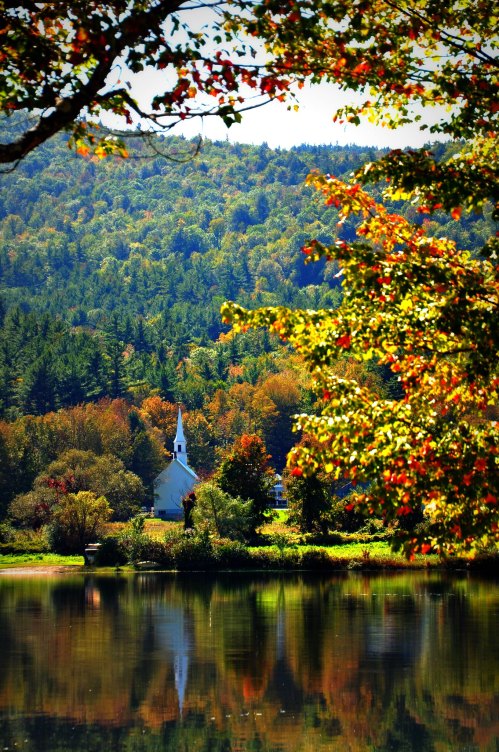 A serene landscape featuring a white church by a lake, surrounded by vibrant autumn foliage and rolling hills.