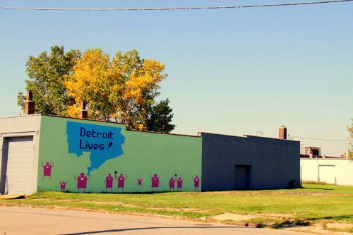Colorful mural on a building in Detroit, featuring the text "Detroit Lives!" and vibrant figures with raised hands.